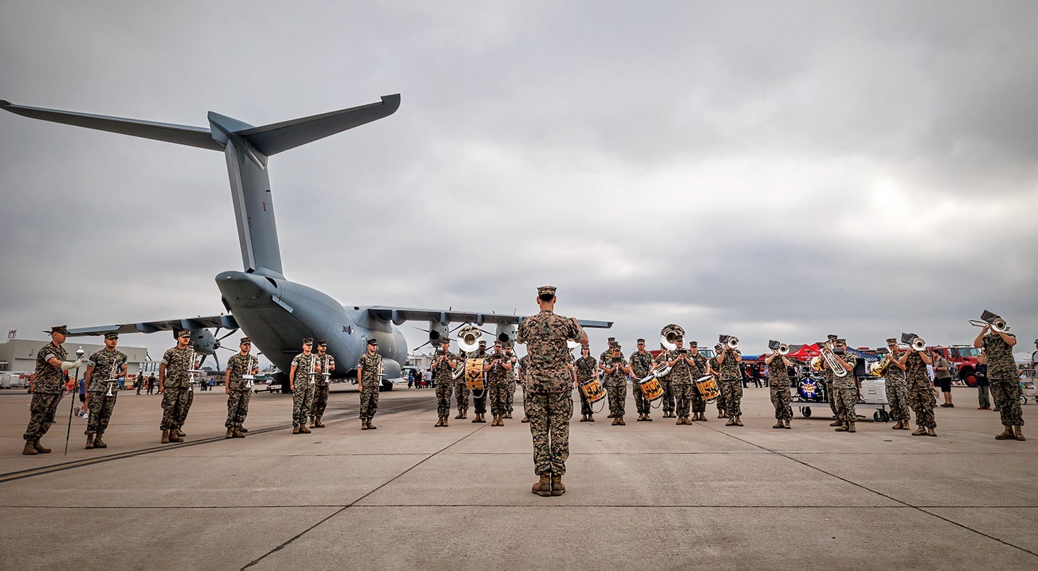 A group of soldiers stands on a tarmac, with a military plane visible in the background.